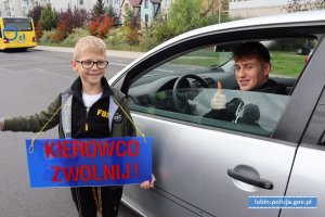 Chłopiec stoi obok samochodu z tabliczką na której napisane jest kierowco zwolnij. W samochodzie mężczyzna uśmiecha się i ma podniesiony kciuk do góry