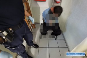 Zatrzymany mężczyzna siedzi na krześle. Obok stoi policjant