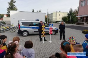 Policjanci ze znakami drogowymi, dzieci w półkolu, w tle radiowóz