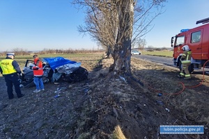 Miejsce wypadku śmiertelnego. Uszkodzony samochód przy drzewie. W tle wóz strażacki policjant, prokurator i biegły