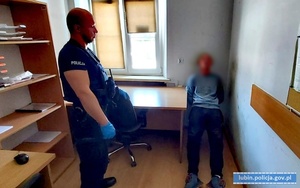 zatrzymany siedzi na krześle w pomieszczeniu z rękoma z tyłu. Obok niego stoi umundurowany policjant