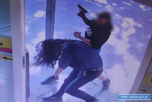 mężczyzna ubrany na ciemno z bronią w ręku szarpie się z osobą. Widok z kamery monitoringu.