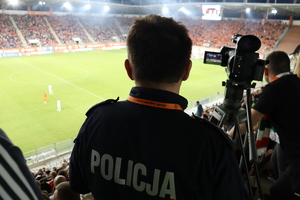Na pierwszym planie widać policjanta, który obserwuje mecz na stadionie, w tle widać trybuny lubińskiego stadionu