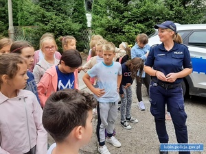 Policjantka rozmawia z dziećmi na temat bezpieczeństwa. W tle radiowóz