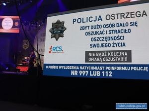 Uroczysta Inauguracja roku akademickiego dla seniorów, policjantka na scenie. Na telebimach wyświetlana ulotka z ostrzeżeniami o oszustwach