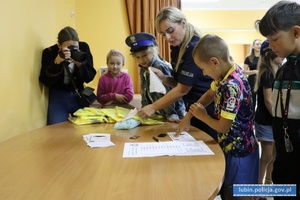 Dzieci rozwiązują krzyżówkę i odbijają palce, żeby zobaczyć linie papilarne oraz dostają kamizelki odblaskowe od policjantki