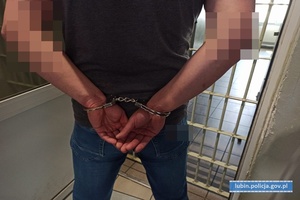 Zatrzymany stoi przed kratą z kajdankami zapiętymi na ręce trzymane z tyłu