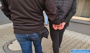 dwóch mężczyzn idących z czego jeden ma założone kajdanki na ręce trzymane z tyłu