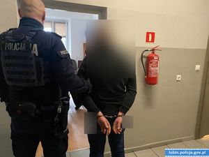 Policjant stoi z zatrzymanym, który ma kajdanki założone na ręce trzymane z tyłu