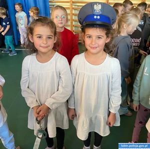 dwie dziewczynki bliźniaczki  w jasnych sukienkach jedna ma na głowię czapkę policyjną, a druga trzyma w reku kajdanki