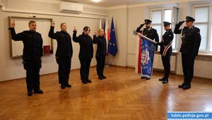 Na zdjęciu czterech policjantów w pozycji na baczność z uniesiona lewa ręką. Przy oknie stoi trzech policjantów z pocztem sztandarowy komendy