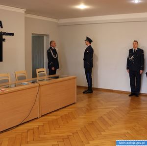 na zdjęciu Komendant Powiatowy Policji w Lubinie wszedł do pomieszczenia gdzie meldunek składa mu drugi policjant. trzeci policjant stoi przy ścianie w pozycji na baczność.