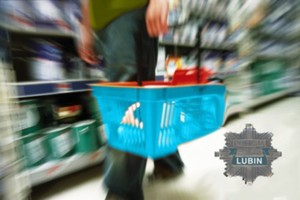 Na zdjęciu widać nogi osoby która idzie sklepem przy rozmazanych regałach sklepowych w ręku trzyma koszyk sklepowy.