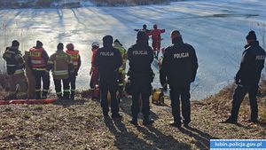 ćwiczenia na zamarzniętym zbiorniku wodnym . Stojący  tyłem policjanci i strażacy przyglądają się czynnościom  ratowniczym prowadzonym na zamarzniętym zbiorniku wodnym