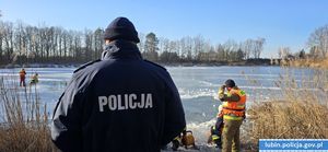 stojący tyłem policjant obserwuje działania prowadzone przez straż na zamarzniętym zbiorniku wody