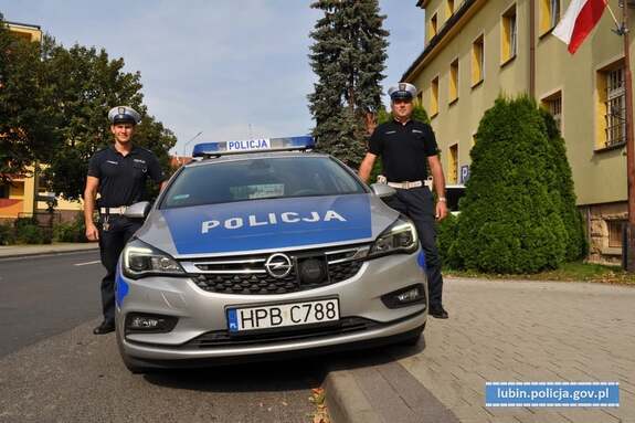 Policjanci ruchu drogowego stoją po lewej i prawej stronie radiowozu przed budynkiem Komendy Powiatowej Policji w Lubinie
