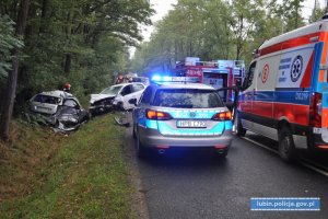 Miejsce wypadku drogowego. Na miejscu radiowóz wóz strażacki. W tle widać dwa uszkodzone samochody osobowe i strażaków