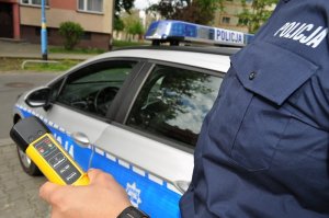 Policjant trzyma w ręku urządzenie do badania stanu trzeźwości. W tle radiowóz