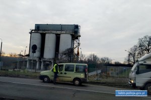 policjant z ruchu drogowego w kamizelce odblaskowej, rękawiczkach lateksowych, i maseczce stoi na jezdni przy zielonym samochodzie. Za nimi w kolejce stoi biały samochód ciężarowy.