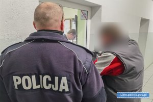 policjant w mundurze stoi tyłem po lewej stronie. Przed nim mężczyzna w maseczce stoi przy okienku dyżurnego i oczekuje na badanie alkomatem. Wizerunek zatrzymanego zamazany.