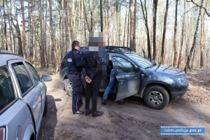 Policjant stoi z zatrzymanym, drugi policjant przeszukuje auto