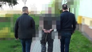 Policjanci operacyjni prowadzą zatrzymanego, który ma ręce zakute w kajdanki zespolone