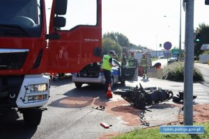 Miejsce wypadku drogowego. Na miejscu leżący motocykl, straż pożarna, policja, karetka pogotowia