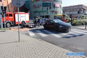 Wypadek drogowy. W tle pojazdy biorące udział w zdarzeniu - samochód osobowy i motocykl, a także służby ratunkowe