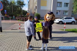 Policjantka przekazuje dziecku kamizelkę odblaskową dziecku, Obok nich stoi komisarz Lew - maskotka dolnośląskiej Policji